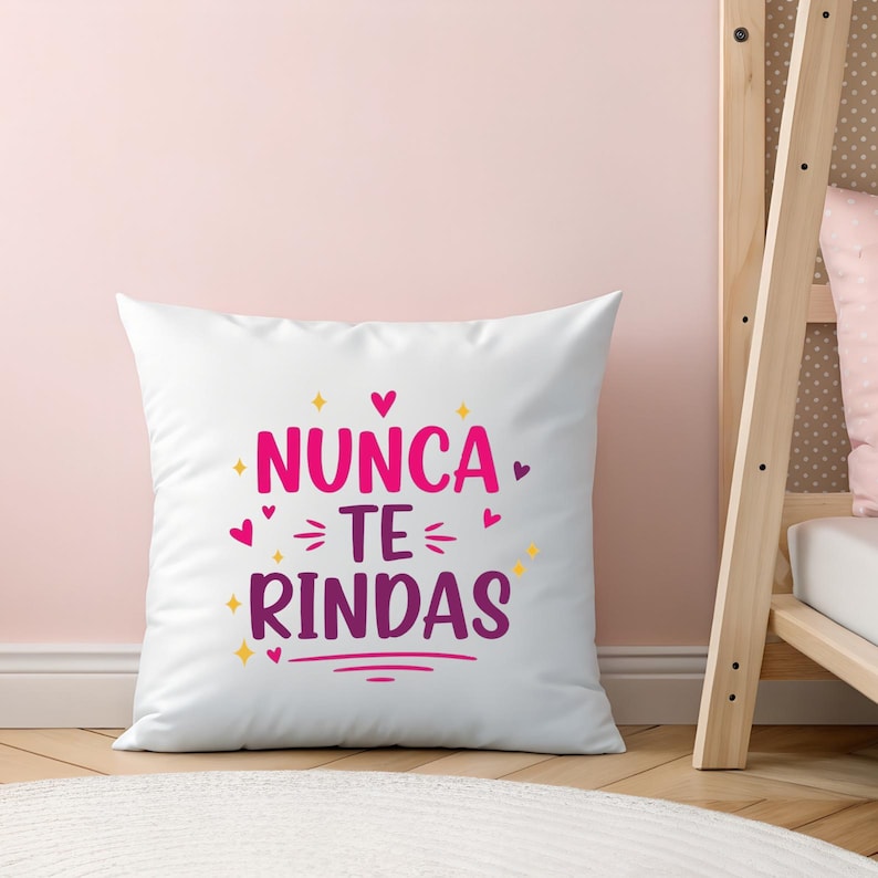 May include: White square pillow with the Spanish text "Nunca te rindas" in pink and purple. The pillow features heart and star accents. The pillow is on a light pink background.