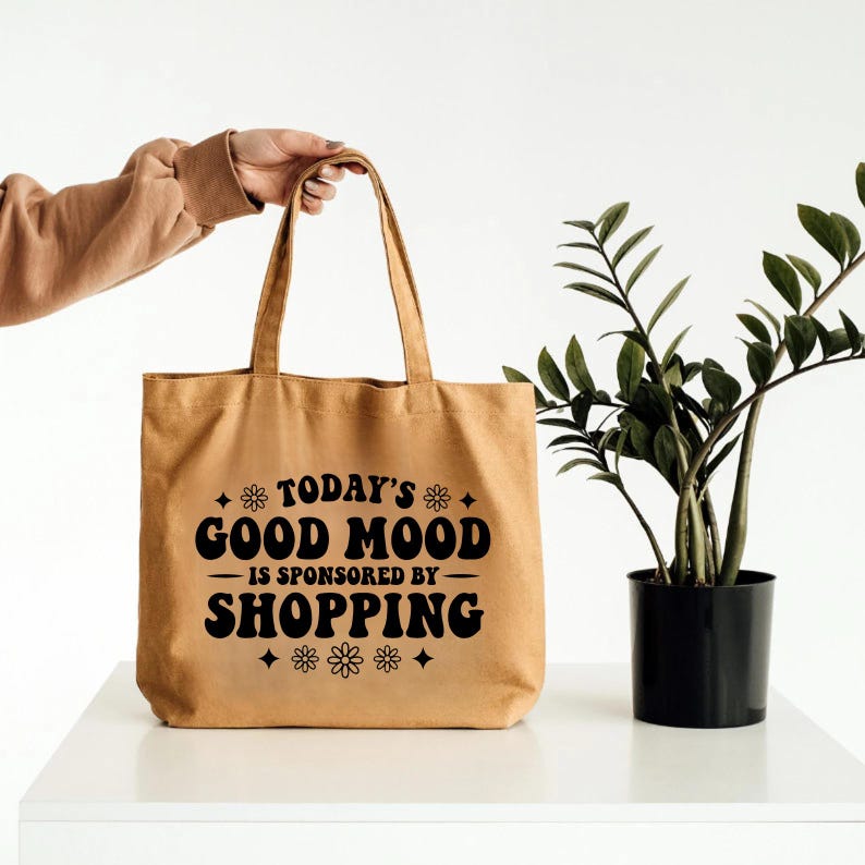 K&ouml;nnte beinhalten: Beige Tragetasche mit schwarzem Text "TODAY'S GOOD MOOD IS SPONSORED BY SHOPPING". Die Tasche wird von einer Person gehalten. Eine Topfpflanze befindet sich rechts von der Tasche.