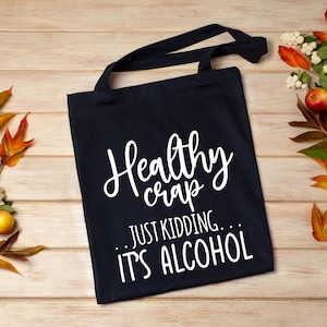 K&ouml;nnte beinhalten: Eine marineblaue Tragetasche mit schwarzen Griffen. Die Tasche hat wei&szlig;en Schreibschrift-Text mit der Aufschrift "Healthy crap... JUST KIDDING... IT'S ALCOHOL". Die Tasche besteht aus strapazierf&auml;higem Stoff und eignet sich zum Tragen von Gegenst&auml;nden.