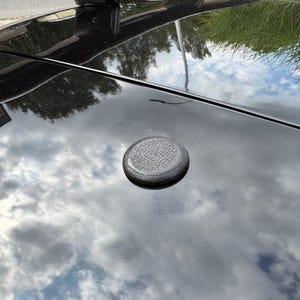 Puede incluir: Un techo de coche negro con un tapón circular texturizado. El techo refleja un cielo nublado y la vegetación circundante. Un alerón negro es visible en la parte superior de la imagen.