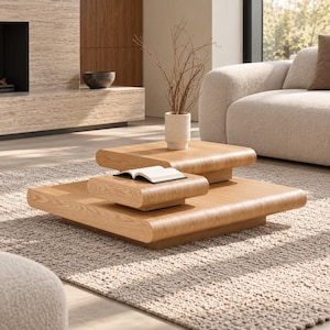 May include: A set of two light-toned wooden coffee tables with rounded edges and a low-profile design. The tables are stacked in a tiered arrangement, with a small white vase containing thin brown branches and an open book placed on top.