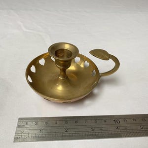 May include: A brass candle holder with a round base and a handle. The base has heart-shaped cutouts. A ruler is placed next to the candle holder, showing measurements in centimetres.