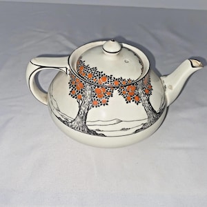 May include: Cream ceramic teapot with a lid, handle, and spout. The teapot is decorated with black tree illustrations and orange fruit. The design wraps around the body and lid. The teapot is on a white surface. A classic tea accessory.