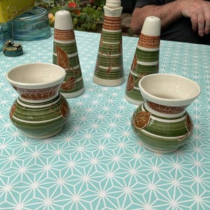 May include: A set of ceramic decorative items, comprising four tall, conical pieces and two small, round vases. The pieces have cream-coloured tops, brown and green stripes, and leaf designs. They are arranged on a light blue patterned tablecloth.