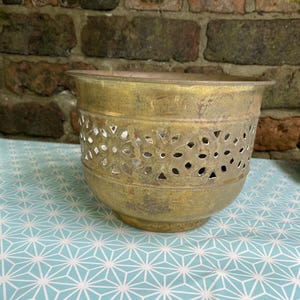 May include: A vintage brass planter with a decorative floral cut-out design around the middle. The planter has a rounded base and a slightly flared rim. The brass has a weathered patina, giving it a rustic appearance. The planter sits on a light blue and white geometric patterned surface.