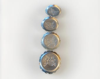 May include: Four small, silver-colored metal dishes arranged vertically. Each dish has a scalloped edge and a raised floral design in the center. The dishes vary slightly in size, with the smallest at the top.