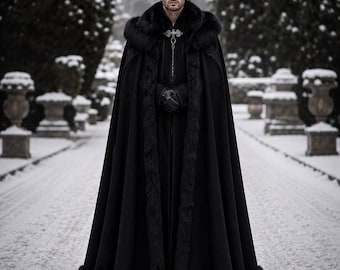 May include: A floor-length black cloak featuring a thick faux fur trim along the hood and hem. The garment includes a decorative silver-toned metal clasp at the neckline and is worn with black gloves, creating a structured and layered appearance.
