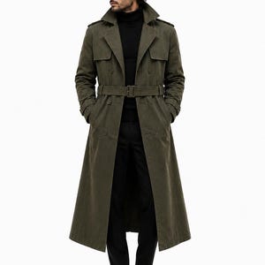 May include: An olive green double-breasted trench coat featuring a wide collar, shoulder epaulets, and a matching waist belt with a buckle. The coat is worn over a black turtleneck sweater and black trousers, paired with black leather boots.