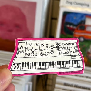 Puede incluir: Una pegatina blanca y negra de un sintetizador con borde rosa. La pegatina presenta una ilustración detallada de las teclas, perillas y botones del instrumento. La dirección del sitio web "JODILYNNDOODLES.COM" está impresa en la esquina inferior derecha.