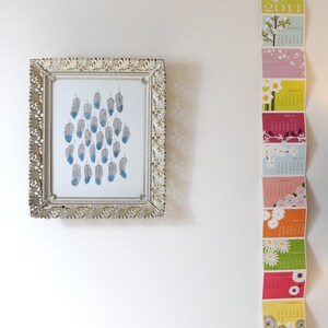 Puede incluir: Una impresi&oacute;n enmarcada de un dise&ntilde;o de plumas azules y grises cuelga de una pared blanca junto a una tira de calendario colorida con el a&ntilde;o 2011 impreso en ella.