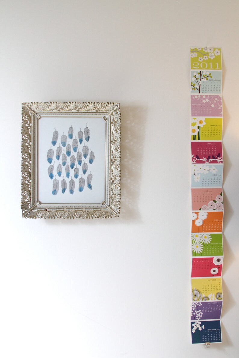 Puede incluir: Una impresi&oacute;n enmarcada de un dise&ntilde;o de plumas azules y grises cuelga de una pared blanca junto a una tira de calendario colorida con el a&ntilde;o 2011 impreso en ella.