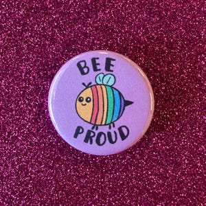 May include: A round, purple button with the words "BEE PROUD" in white. The button features a cartoon bee with a rainbow-colored body and a smiling face. The background is a sparkly, magenta surface.