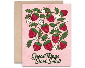 May include: A greeting card with a pink background, featuring a strawberry illustration. The strawberries are red with green leaves and white flowers. The text "Great Things Start Small" is in green script.