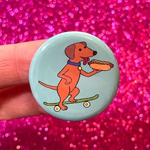 May include: A blue and white button featuring a cartoon dog on a skateboard holding a hot dog. The dog is orange with a red tongue sticking out.
