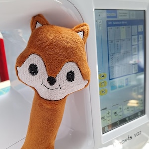 以下が含まれることがあります： ミシンハンドルカバー、キツネの形をしたぬいぐるみ。 カバーは主に茶色で、白い顔、黒い目、笑顔の口があります。 ミシンは白です。