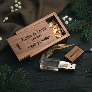 Op de afbeelding: Een houten USB-stick en doos met de namen "Katie & Louis" en de datum "12-2-2023" gegraveerd. De doos is open en gevuld met gedroogde bloemen. De USB-stick is zilverkleurig en transparant.