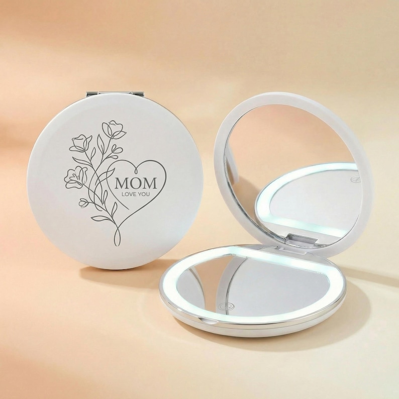 May include: A round white compact mirror with an LED light feature. The closed mirror displays a black line art illustration of a floral bouquet and a heart shape containing the text MOM LOVE YOU. The open mirror shows a circular reflective surface.