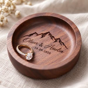 Op de afbeelding: Een ronde donkerbruine houten ringhouder met een in laser gegraveerde berggrafiek boven de tekst Oliver & Amelia en de datum 14.09.2026. Een goudkleurige ring met een heldere ronde edelsteen ligt in de cirkelvormige houten schaal.