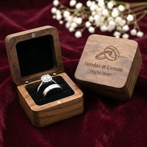 May include: A wooden ring box with a silver engagement ring and wedding band. The box is open, revealing a black velvet interior. The closed box has an engraved design of two rings and the names "Sundas & Usman" with the date "03/11/2017".