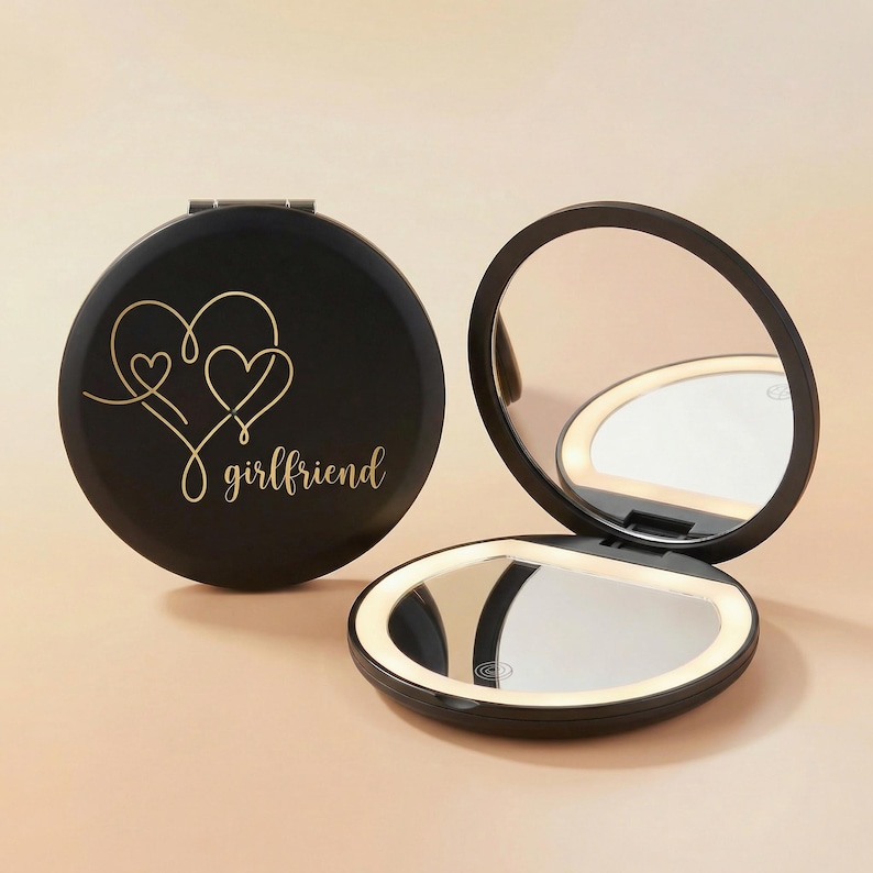 May include: A round black compact mirror with an LED light ring. The closed mirror features a gold-colored line art design of two connected hearts and the word "girlfriend" in cursive script. The open mirror shows a reflective surface with a light.