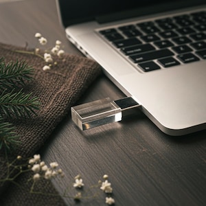 May include: A clear, rectangular USB flash drive inserted into a silver laptop. The drive is made of transparent material. The laptop sits on a dark wooden surface, next to a brown cloth and sprigs of white flowers and green pine needles.