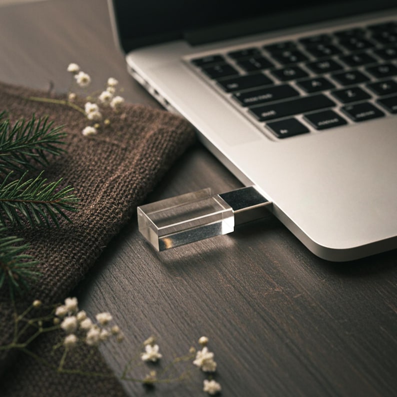 May include: A clear, rectangular USB flash drive inserted into a silver laptop. The drive is made of transparent material. The laptop sits on a dark wooden surface, next to a brown cloth and sprigs of white flowers and green pine needles.