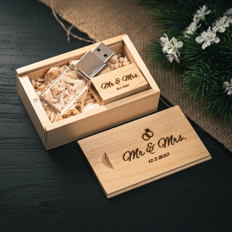 May include: A wooden USB flash drive and a small wooden card with the inscription "Mr. & Mrs." and the date 12.2.2023. The USB drive is in a wooden box filled with wood shavings. The box is open, and the card is next to it.