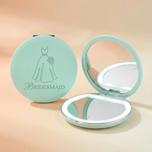 May include: A round, mint green compact mirror with a hinged lid. The lid features a line art illustration of a dress and the text "BRIDESMAID" in a script font. The open mirror displays a circular LED light ring around the reflective surface.