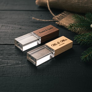 May include: Two wooden USB flash drives with clear acrylic bodies. One is dark brown with "Vaster & Solin" engraved, the other is light brown with "Mr. & Mrs." and a date. The drives are on a dark wooden surface.