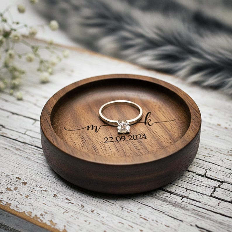 Puede incluir: Un plato redondo de madera oscura para anillos con un anillo de oro con una gema transparente. El plato est&aacute; grabado con las iniciales "m k" y la fecha "22.09.2024". El plato para anillos est&aacute; sobre una superficie de madera blanca envejecida.