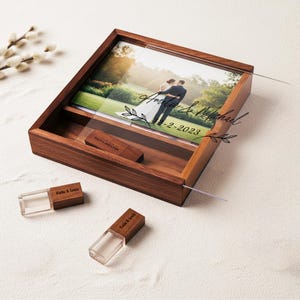 Pode incluir: Uma caixa de fotos de madeira com uma tampa deslizante transparente, contendo uma foto de casamento e uma unidade USB. A caixa de fotos tem os nomes "Amelia & Michael" e a data "2-2-2023" gravados. Duas unidades USB adicionais são visíveis.