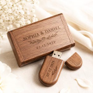 Puede incluir: Una caja rectangular de madera marrón oscuro para memoria USB grabada con el texto SOPHIA & DANIEL, un corazón, ramas de hojas y la fecha 07.12.2025. Al lado hay una memoria USB de madera a juego con tapa extraíble, también grabada 07.12.2025.