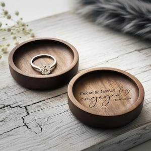 Puede incluir: Dos cuencos redondos de madera oscura. Uno contiene un anillo de compromiso plateado con una piedra transparente. El otro est&aacute; grabado con las palabras "Oscar & Jessica engaged 22.09.2024" y un coraz&oacute;n. Sobre una superficie de madera blanca.