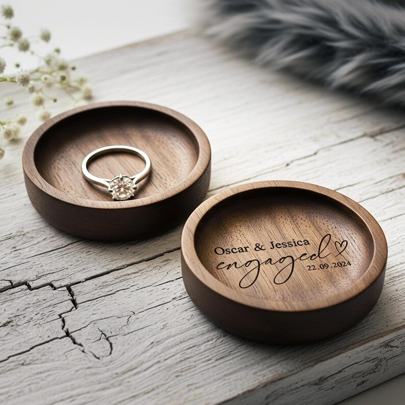 Puede incluir: Dos cuencos redondos de madera oscura. Uno contiene un anillo de compromiso plateado con una piedra transparente. El otro est&aacute; grabado con las palabras "Oscar & Jessica engaged 22.09.2024" y un coraz&oacute;n. Sobre una superficie de madera blanca.