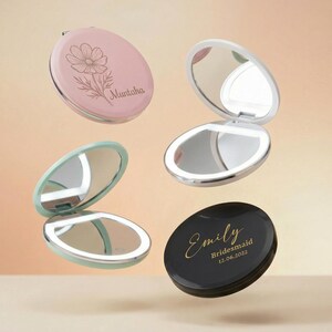 May include: Four round compact mirrors with LED lights. One is pink with a floral line drawing and the name Muntaha. One is black with gold text reading Emily Bridesmaid 12.06.2022. Two are open, showing reflective surfaces and white light rings.
