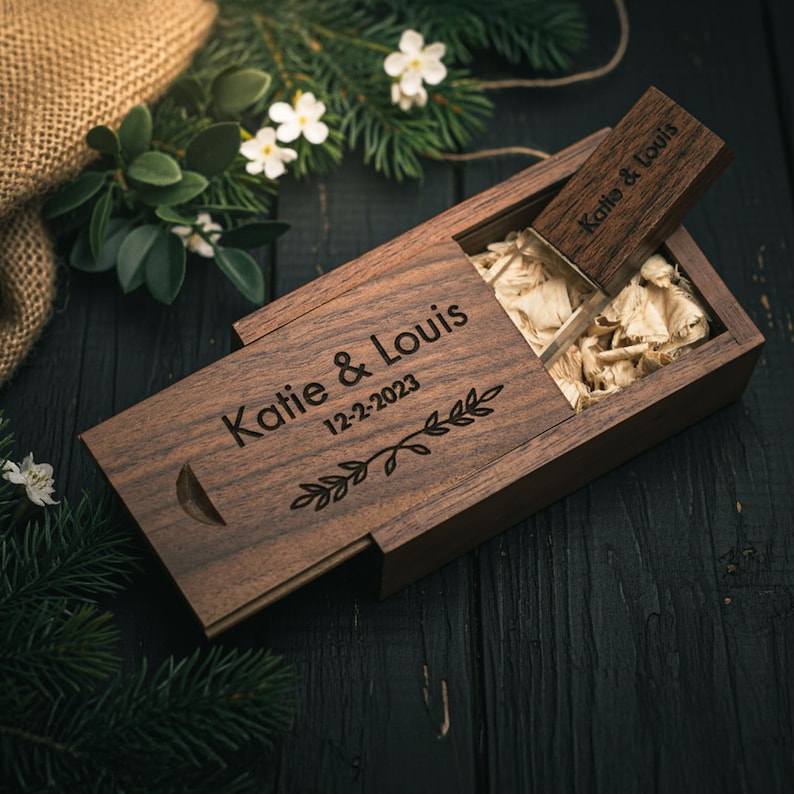May include: A wooden USB flash drive and case set. The case is engraved with the names "Katie & Louis" and the date "12-2-2023", along with a decorative leaf design. The USB drive is partially inserted into the case, which is filled with shredded paper.