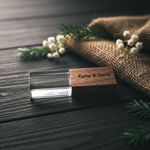 May include: A personalized USB flash drive with a clear acrylic body and a wooden cap engraved with "Katie & Louis." The flash drive is displayed on a dark wooden surface, with decorative greenery and burlap in the background.