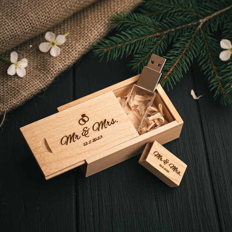 May include: A wooden USB flash drive in a light-colored wooden box. The box is engraved with "Mr & Mrs" and the date 12.2.2023. The USB drive is partially visible, and the box is open, revealing wood shavings inside. A small matching wooden tag is also present.