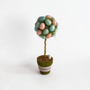 Puede incluir: Un topiario decorativo de Pascua con un grupo de huevos rosas y verde azulados brillantes. Los huevos están dispuestos en forma esférica sobre un tallo delgado y marrón. El tallo está plantado en una maceta verde con una banda de encaje blanco y una pequeña figura de conejito blanco.