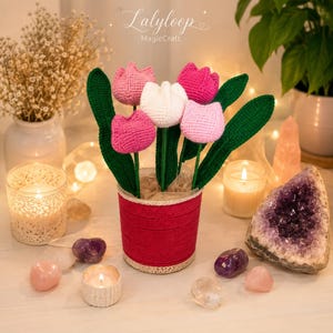 Può includere: Un bouquet di tulipani all'uncinetto con quattro fiori nelle tonalità del rosa e del bianco con steli e foglie verdi, sistemato in un vaso cilindrico rosso con un bordo in pizzo color crema alla base. Il testo Lalypoop MagicCraft è visibile.