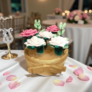 Może przedstawiać: Ręcznie robiona kompozycja kwiatowa z szydełkowymi różami w kolorze różowym i kremowym, zielonymi liśćmi i złotym koszem z kokardą. Kompozycja stoi na białym obrusie, z rozrzuconymi różowymi płatkami róż.