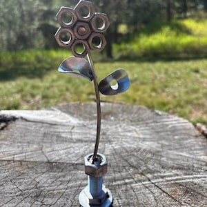 Peut inclure: Sculpture florale en métal faite à la main. La tête de la fleur est composée de plusieurs écrous hexagonaux disposés en cercle. La tige et les feuilles sont en métal. La sculpture est montée sur une base boulon et écrou, sur une souche de bois.