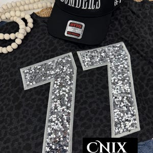 May include: A black baseball cap with the word BOMBERS and #71 embroidered in white, paired with a dark gray leopard print t-shirt featuring large silver sequined number 77 patches. The cap has an Otto brand sticker on the front brim.