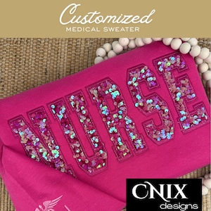 May include: A bright pink sweater featuring the word NURSE in large, block-style letters filled with iridescent, multi-colored sequins. The letters are outlined in matching pink thread, and the fabric of the sweater is a solid, vibrant pink color.