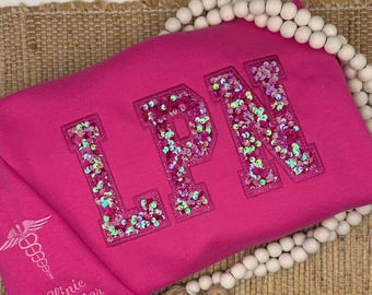 以下が含まれることがあります： 鮮やかなピンク色のスウェットシャツで、LPNの文字が虹色に輝くピンクと白のスパンコールで埋め尽くされています。袖には白いカドゥケウスのシンボルとClinic Directorという文字が刺繍されています。白い木製ビーズのネックレスが添えられています。