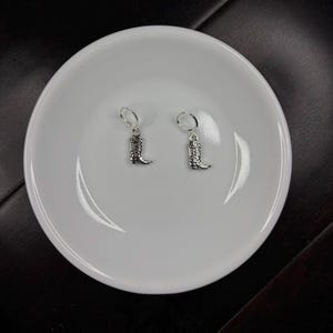 May include: A pair of silver-colored cowboy boot earrings with lever-back closures. The earrings are displayed on a white, round plate against a dark background. The boots have detailed designs.