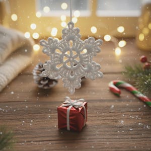 Könnte beinhalten: Ein weißer, gehäkelter Schneeflocken-Ornament hängt über einer kleinen, roten Geschenkbox, die mit einem weißen Band gebunden ist. Eine Zuckerstange und ein Tannenzapfen befinden sich ebenfalls auf der Holzoberfläche, mit sanften Lichtern im Hintergrund.