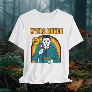 Könnte beinhalten: Weißes T-Shirt mit einer Grafik von Michael Myers. Das Design enthält den Text "Myers Munch" über einem Regenbogen und einer Schüssel mit Müsli. Das Shirt hat auch den Text "Free Mask Inside!"