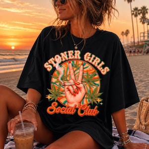 Op de afbeelding: Een zwart T-shirt met korte mouwen en een grafische print met de tekst "STONER GIRLS Social Club". Het ontwerp toont een hand die een vredesteken maakt, een brandende sigaret en cannabisbladeren tegen een cirkelvormige oranje en groene achtergrond.