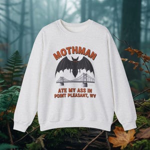 May include: A light grey sweatshirt featuring a graphic of Mothman with a bridge and the text "MOTHMAN ATE MY ASS IN POINT PLEASANT, WV" in orange and white. The design includes a black bat-like creature with red eyes.